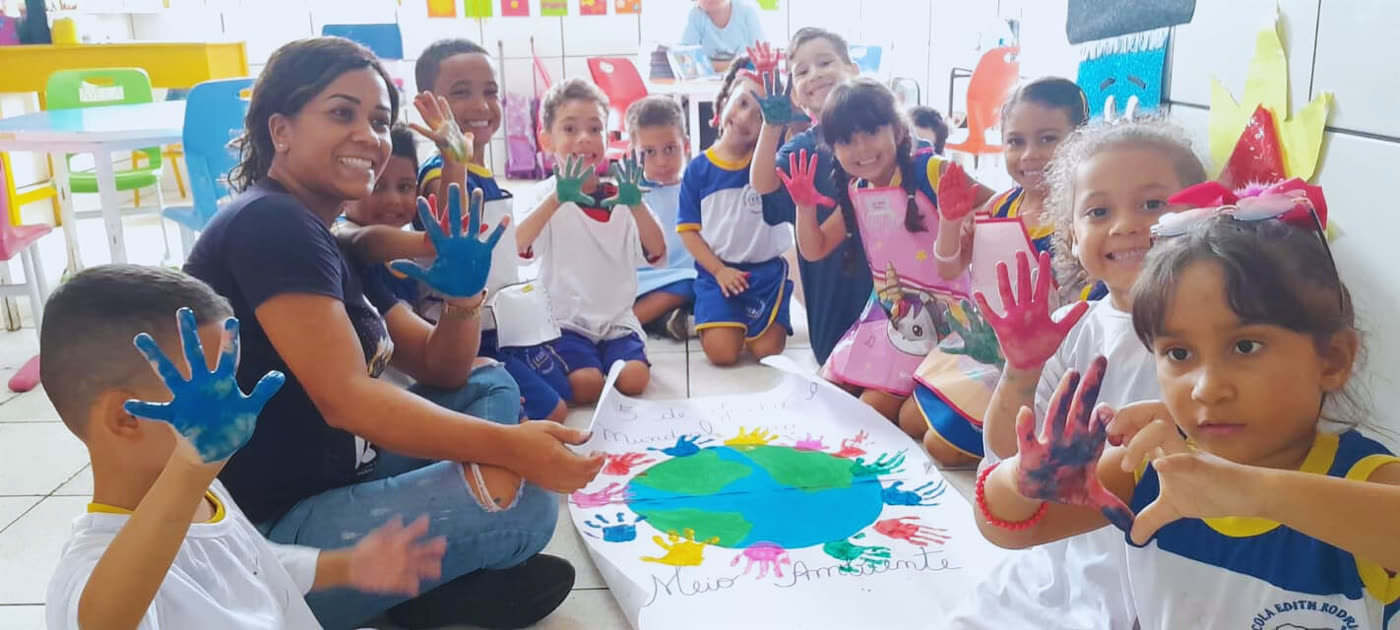 Professora e crianças da Educação Infantil com as mãos pintadas em volta de um trabalho coletivo