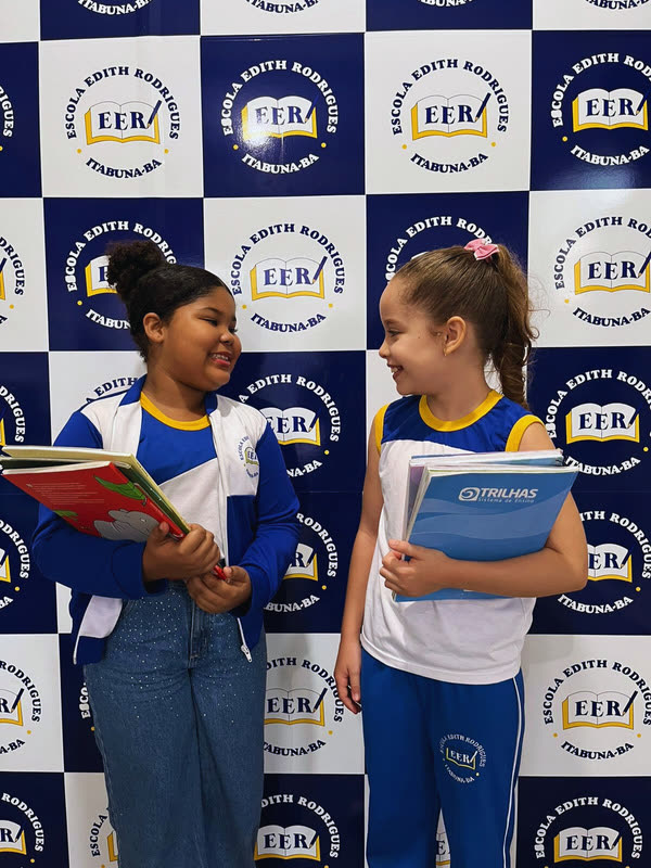Duas alunas do Fundamental I com livros diante do painel da escola